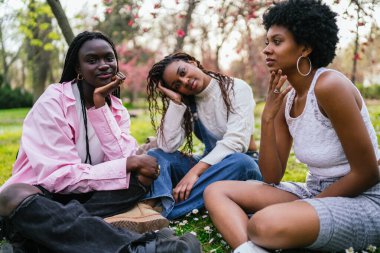 Üç genç Afrikalı kadın düşünceli bir şekilde parkta çimlerin üzerinde oturur ve çiçeklerle çevrili kameraya bakarlar..