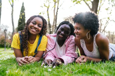 Üç Afrikalı genç kadın çimlere uzanmış, gülümsüyor ve arkadaşlıklarından keyif alıyorlar. Mutluluk ve dostluk kavramı