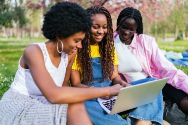 Üç neşeli Afrikalı kadın çimenlerde yan yana oturur parktaki ağaçların altında dizüstü bilgisayarda çalışırlar..