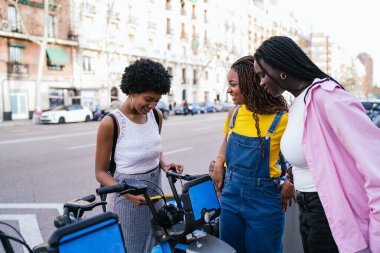 Üç Afrikalı kadın şehrin caddesinde elektrikli bisiklet kiralamak için akıllı telefon kullanıyor, gülümsüyor ve anın tadını çıkarıyorlar..