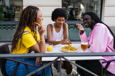 Üç Afrikalı kadın, şehir meyhanesinin terasında nachos yiyip içmekten zevk alarak neşeli ve mutlu bir atmosfer yaratıyor..