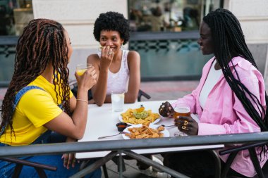 Üç Afrikalı kadın açık hava taverna masasında nachos ve içki paylaşıyor, gülümsüyor ve birlikte vakit geçiriyorlar..