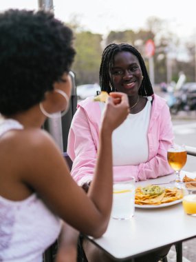 İki Afrikalı kadın, açık hava tavernasında cips ve içkiden hoşlanırlar. Hoş bir sohbete girerler..