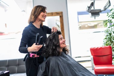 Bir kuaför, parlak ve modern bir kuaförde mutlu bir müşterinin saçını şekillendirir. Etkileşim neşeli ve profesyonelce. Neşeli salon deneyimi kavramı.