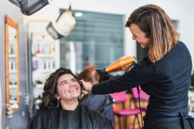 Kuaför, gülümseyen bir müşterinin saçını kuaförde saç kurutma makinesiyle şekillendirir. Neşeli ve arkadaş canlısı saç stili deneyimi kavramı.