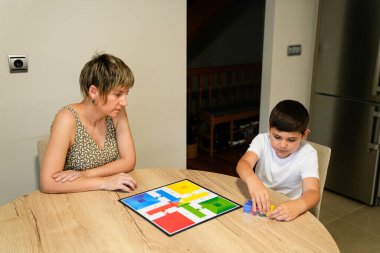 Bir anne, oğlunu yemek masasında Parcheesi oynarken dikkatle izler. Sahne, onların odaklanmış etkileşimlerini ve ortak zevklerini vurguluyor. Aile bağları kavramı.