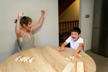 Bir anne masa başı sopa oyununda zaferi sevinçle kutlar. Oğlu heyecanı paylaşırken kollarını zaferle kaldırır. Aile eğlencesi ve başarı kavramı.