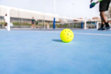 Sarı bir picleball topu, arka planda bulanık bir grup oyuncuyla mavi bir kortta duruyor. Oyuna hazırlık ve çeşitlilik kavramı.