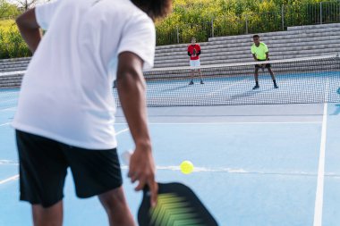 Çok ırklı rakipleri hazır beklerken, Afrika kökenli Amerikalı bir picleball oyuncusu mavi sahada topa servis yapıyor. Rekabet ve hazırlık kavramı.