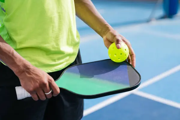 Yakın plan çekimde, bir oyuncunun elinde bir picleball raketi ve topu, oynamaya hazır bir şekilde tutar. Hazırlık ve odaklanma kavramı.