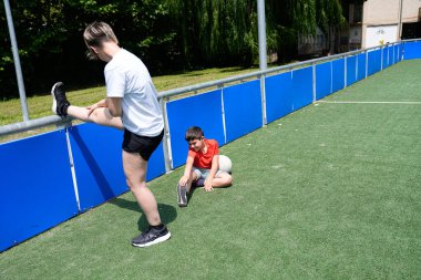 Anne ve oğlu maçtan önce mavi bariyerlerle bir futbol sahasında esniyorlar. Zindelik, bağlılık ve spor hazırlığı kavramı.