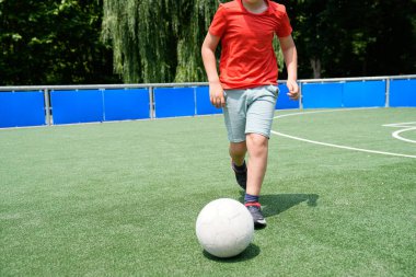 Bir çocuk futbol maçı sırasında topu kontrol eder, güneşli bir sahada ilerlerken adımlarına odaklanır. Oynayarak öğrenme kavramı.