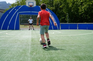 Genç bir çocuk kaleye doğru top sürerken topu kontrol ediyor, oyuna odaklanıyor. Futbol öğrenme ve pratik yapma kavramı.