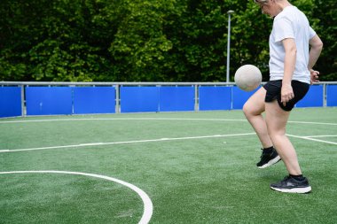 Bir kadın sahada futbol topunu tekmeliyor. Saha yeşil ve mavi, ve kadın beyaz gömlek ve siyah şort giyiyor.