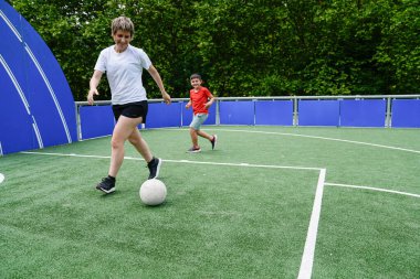 Annesi, oğlu arkasından koşarken top sürüyor ve hızlı bir oyun oynuyor. Aktif aile eğlencesi ve açık havada formda kalma kavramı.