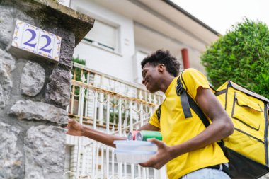 Sarı sırt çantalı bir teslimatçı eve yemek götürürken gülümsüyor. Arkadaş canlısı ev teslimat hizmeti kavramı.