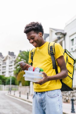 Gülümseyen bir Afrikalı kurye teslimat sırasında elinde yiyecek ve içeceklerle telefonunu kontrol ederken yürüyor. Yaya olarak yiyecek teslimatı kavramı.