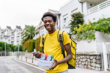 Mutlu bir Afrikalı kurye yiyecek, içecek ve telefonunu meskun bir caddede yürürken saklar. Yaya olarak kentsel gıda teslimatı kavramı.