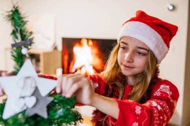 Kız Noel ağacına bir yıldız süsü ekliyor, arkasında da sıcak bir ateş parlıyor. Şenlikli dekorasyon ve sıcaklık kavramı.