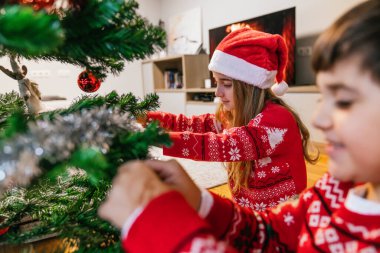 Noel süveteri giyen iki kardeş Noel ağacını dikkatlice süslüyorlar. Kardeş takım çalışması ve festival gelenekleri kavramı.