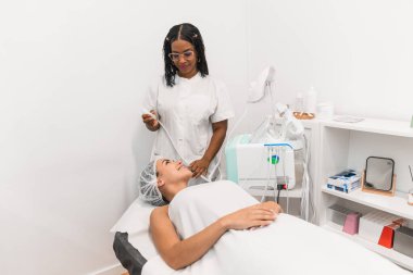 Gülümseyen güzellik uzmanı, profesyonel bir güzellik salonundaki yatakta yatan genç bir kadının yüzüne yakın bir yerde ultrason cihazı tutuyor.