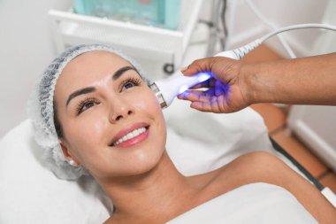 Güzellik uzmanı, güzellik salonundaki gülümseyen bir kadının yüzüne lazer tedavisi uyguluyor.