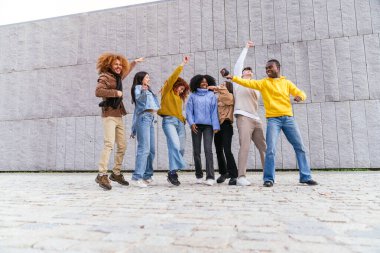 Çok ırklı bir grup arkadaş modern gri bir duvarın önünde zıplıyor ve dans ediyorlar.