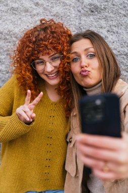 İki genç kadın birlikte selfie çekiyorlar, biri barış işareti yapıyor ve diğeri dudaklarını büzüyor.