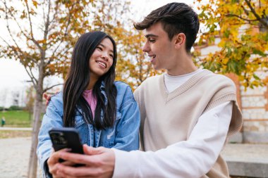 Genç çift parkta bir bankta oturuyor, akıllı telefon kullanıyor ve birbirlerine gülümsüyorlar.