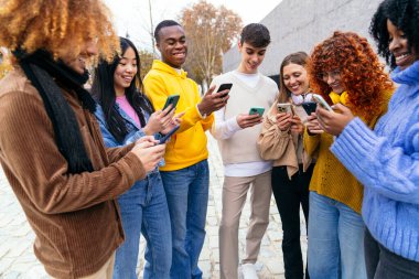 Bir grup çok ırklı arkadaş dışarıda ayakta gülüyor ve akıllı telefonlarını kullanıyorlar.