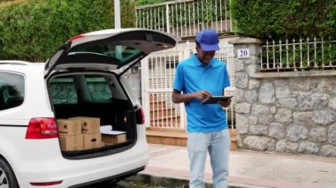 Mavi üniformalı gülümseyen bir teslimatçı arabasının yanında tabletle paketleri kontrol ediyor. Verimli paket izleme ve lojistik hizmetleri kavramı.