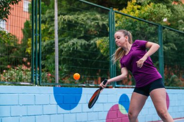 Pickleball oynayan kadın sporcu, renkli bir kortta küreğiyle topa vuruyor.