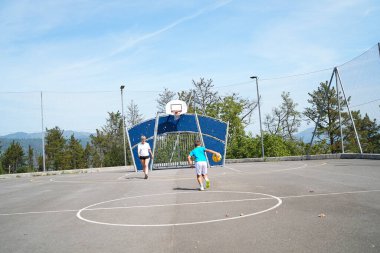 Anne ve oğlu, ağaçlar ve dağlarla çevrili açık bir sahada basketbol oynayarak güneşli bir günün tadını çıkarıyorlar.
