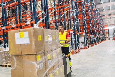 Yüksek görüşlü yelek giyen bir depo işçisi büyük kutuları dağıtım merkezinde palet krikosuna taşıyor, lojistik ve siparişleri düzene sokuyor.