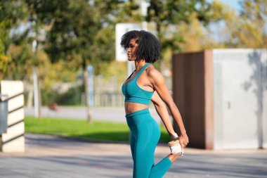 Turkuaz spor giyimli, formalı bir zenci kadın şehir parkında egzersiz yapmadan önce ısınıyor ve bacaklarını esnetiyordu.