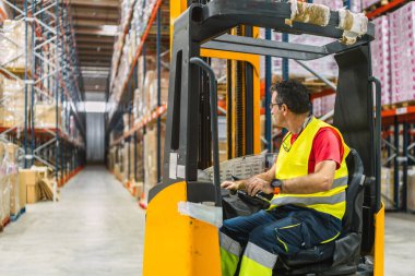 Güvenli yelek giyen konsantre olgun bir adam, lojistik merkezinin geniş ambarındaki rafların arasında forklift kullanıyor.