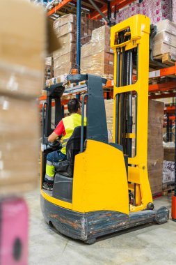 Lojistik işçisi forklift kullanıyor. Büyük bir depoda raflardaki malları düzenliyor.