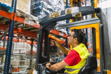 Güvenlik yeleği giyen depo işçisi forklift kullanıyor ve modern lojistik merkezindeki paketleri taramak için barkod okuyucu kullanıyor.