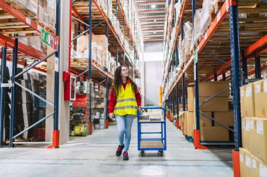 Güvenli yelek giyen genç bir kadın büyük dağıtım merkezindeki rafların arasına el arabası koyuyor.