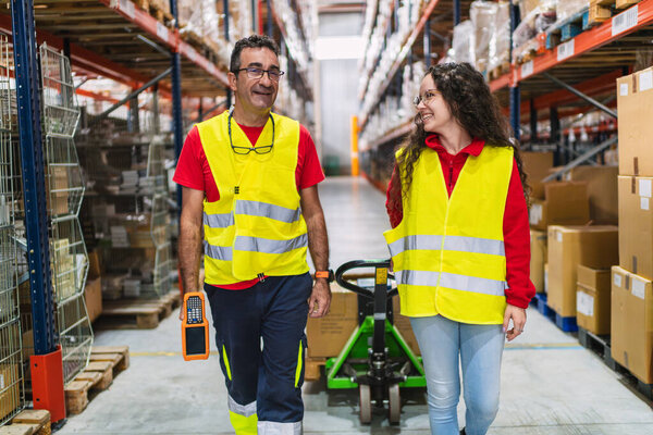 Warehouse workers are walking through the warehouse, using a handheld computer and pulling a pallet jack, smiling and working together