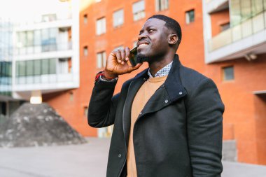 Genç Afrikalı iş adamı, modern bir şehir ortamında dururken akıllı telefonuyla gülümsüyor ve konuşuyor.