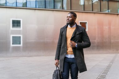 Kendine güvenen genç Afrikalı girişimci modern ofis binasının dışında yürüyor, evrak çantası taşıyor ve başka tarafa bakıyor.