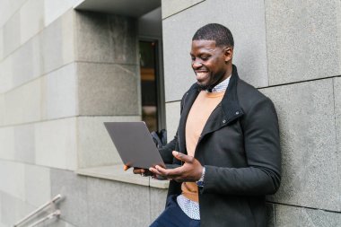 Genç işadamı dizüstü bilgisayar kullanıyor ve modern ofis binasının dışında gülümsüyor, iş hayatı dengesinin keyfini çıkarıyor