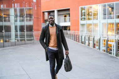 Genç Afrikalı iş adamı modern bir ofis binasının önünde elinde evrak çantası ve gülümsüyor.