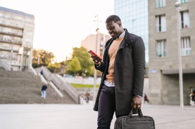 Şık bir işadamı, bir çanta taşırken akıllı bir telefon kullanarak güvenli bir şekilde şehir sokaklarında yürüyor, modern profesyonelliği somutlaştırıyor.