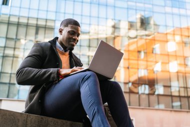 Genç Afrikalı iş adamı, modern bir ofis binasının önünde dizüstü bilgisayarıyla çalışıyor, şehir ortamının tadını çıkarıyor.
