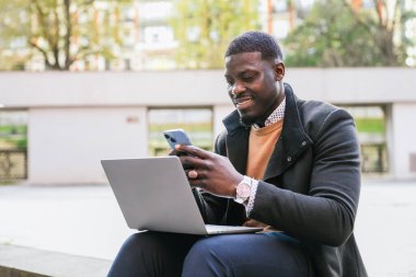 Dışarıda çalışan gülümseyen Afrikalı genç iş adamı, modern bir şehir ortamında iletişim kurmak ve iletişim kurmak için bir laptop ve akıllı telefon kullanıyor.