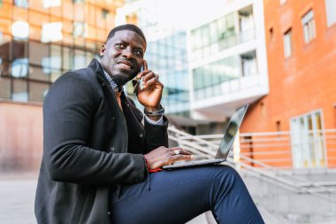 Genç Afrikalı iş adamı modern ofis binalarının önünde oturuyor, dizüstü bilgisayarda çalışıyor ve cep telefonuyla konuşuyor.