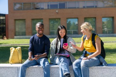 Üç neşeli çoklu etnik üniversite öğrencisi kampüs bahçesindeki duvarda otururken akıllı telefonlar kullanıyorlar.