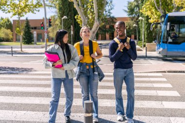 Sırt çantalı üç neşeli çoklu etnik üniversite öğrencisi yaya geçidinde yürüyorlar ve kampüse gidiyorlar.